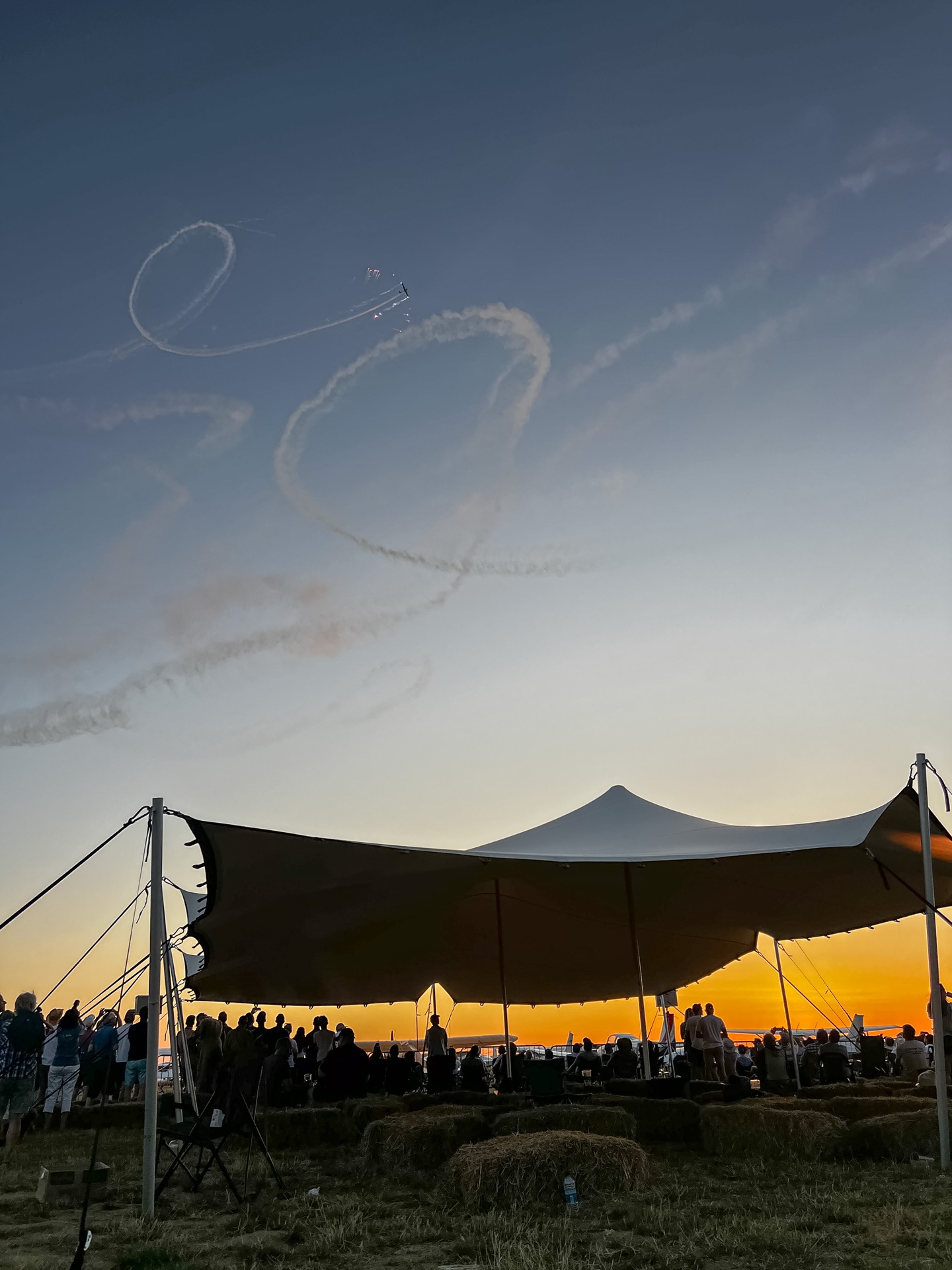 Stretch tent full of spectators at sunset with aerobatic smoke trails overhead